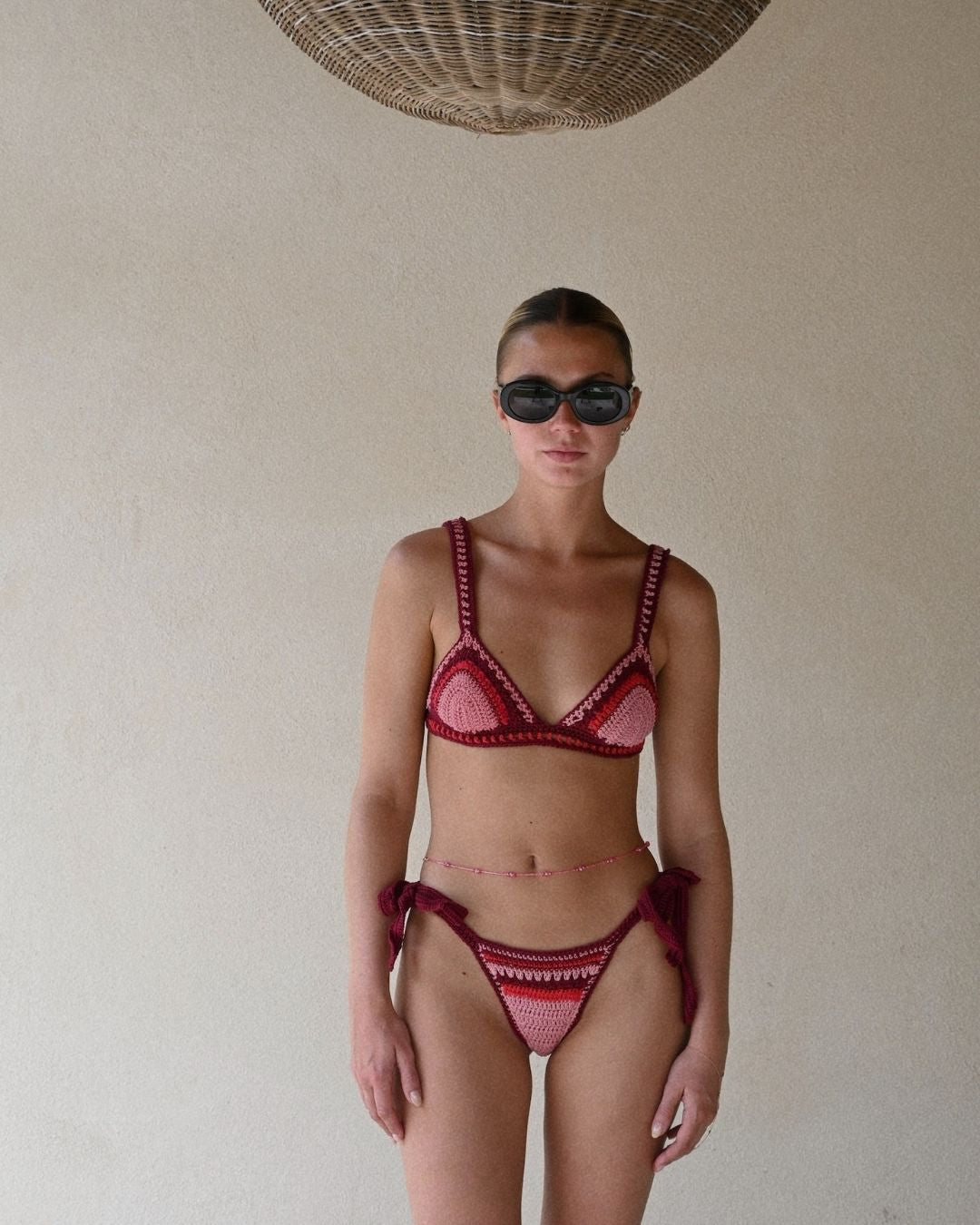 Full front view of a model standing indoors, wearing the vibrant cherry and pink Bianca crochet bikini with large dark sunglasses.
