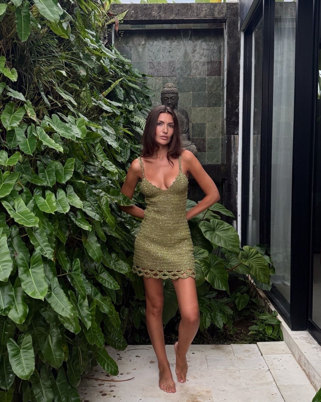Full front view of a model in a tropical garden with a Buddha statue, wearing the radiant olive Apple Pie crochet dress.