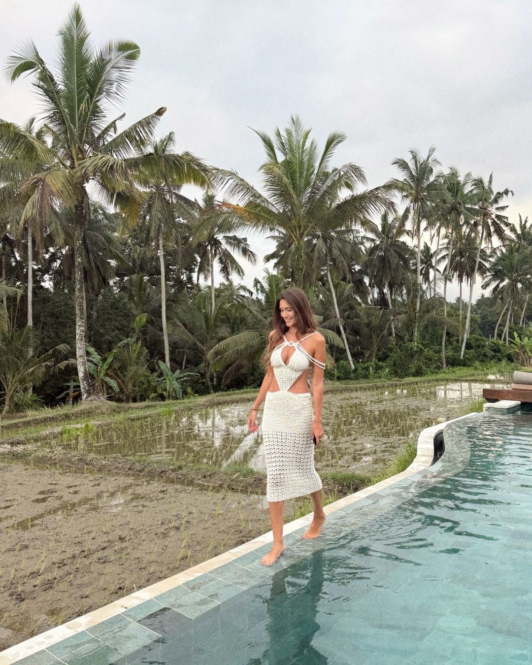 a full-body shot of a model with long brown hair walking barefoot along the edge of an infinity pool. she wears the cream mélissa crochet midi dress with its cut-outs and multi-strap details. the background features lush palm trees, a flooded rice paddy field, and traditional balinese structures under a cloudy sky.