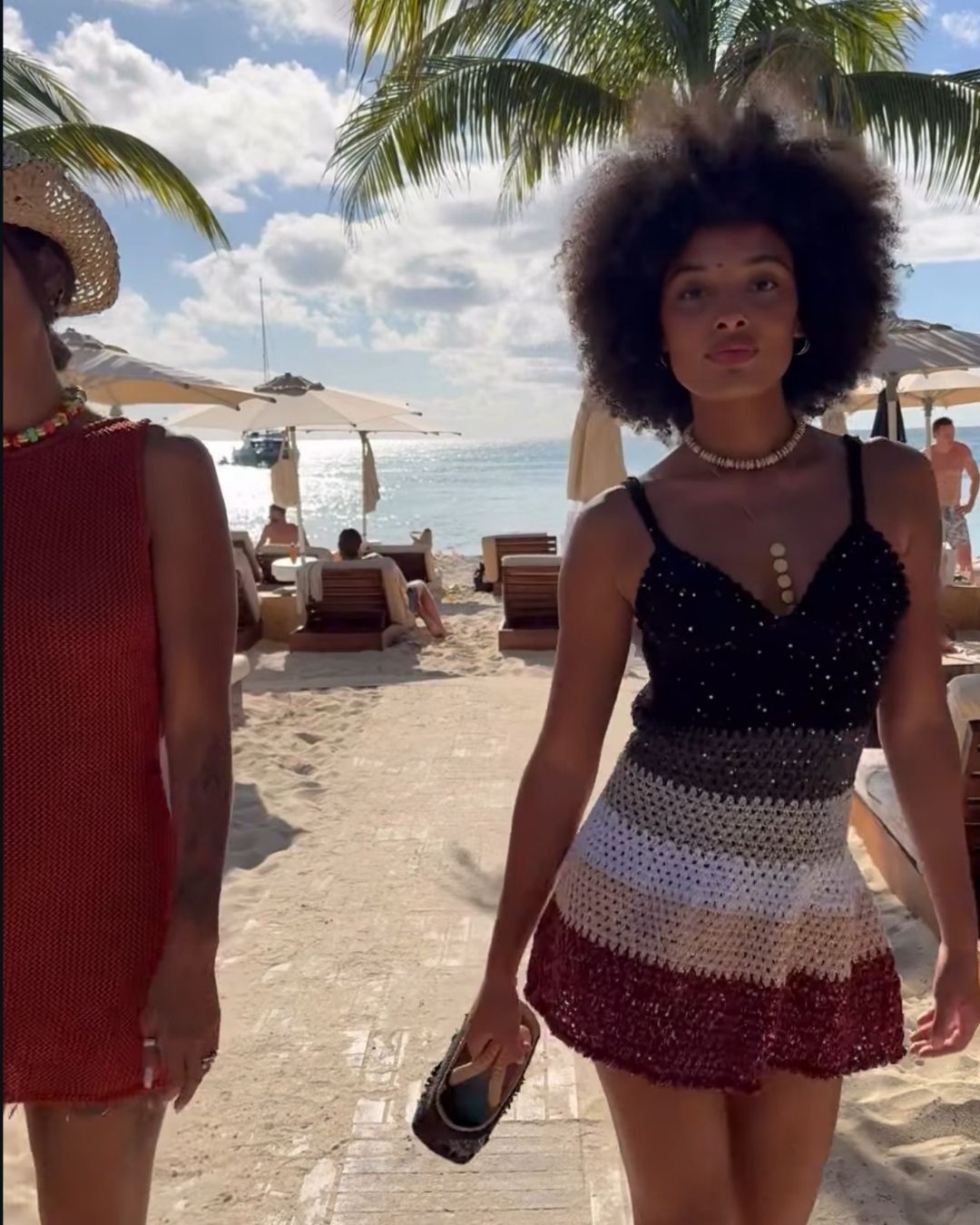 Lifestyle shot of a model wearing the Sienna multi-stripe mini dress. The photo captures the shimmering sequins and the vibrant hand-crocheted gradient of black, grey, white, and burgundy, showcasing the dress's flattering fit and luxury resort aesthetic.